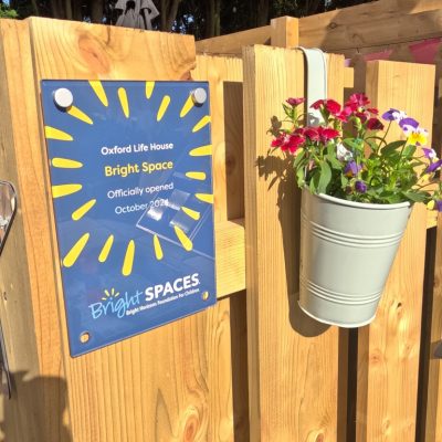 Brightly coloured naming plaque in garden designed by Bright Space with Life Charity for Oxford Life House, on garden fence with hanging bucket of bright flowers