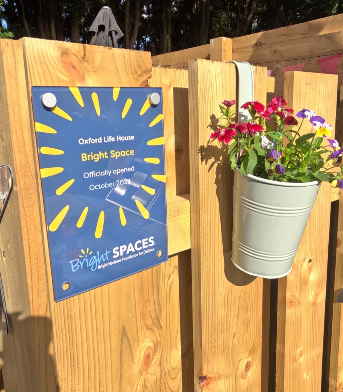 Brightly coloured naming plaque in garden designed by Bright Space with Life Charity for Oxford Life House, on garden fence with hanging bucket of bright flowers